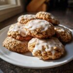 A tempting stack of freshly baked iced oatmeal cookies piled high on a white plate, glistening under natural light.