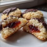 Close-up of Italian butter cookies, one split open revealing a bright red jam center, dipped in dark chocolate.