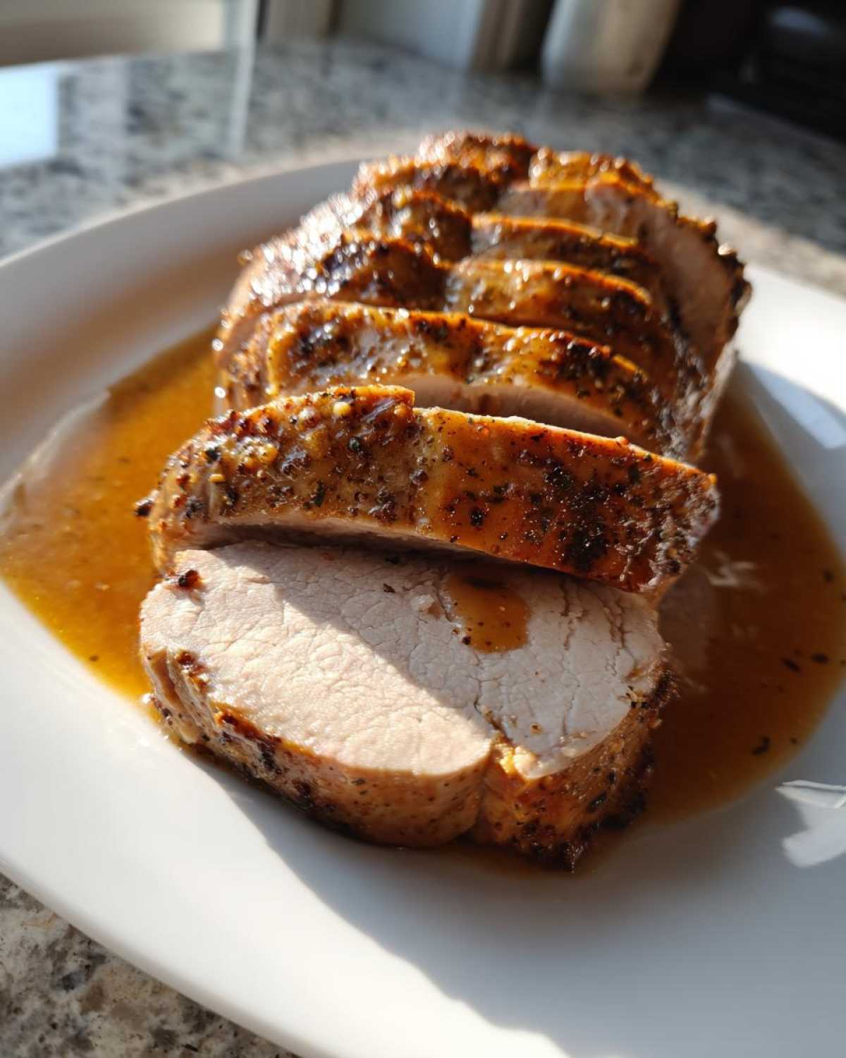 Slices of juicy pork loin roast with a dark, seasoned crust resting in its pan juices on a white plate.