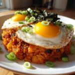 A mound of vibrant kimchi fried rice topped with two sunny-side-up eggs, seaweed, and sliced green onions.