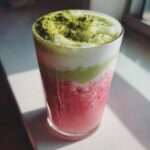 A tall glass showcasing a layered strawberry matcha latte with a bright pink base, white foam, and matcha powder sprinkled on top.