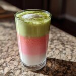A tall glass showing a layered strawberry matcha latte with white milk, pink strawberry layer, and green matcha foam.