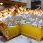 Close-up of a thick slice of lemon meringue pie showing bright yellow curd and tall, torched meringue peaks.