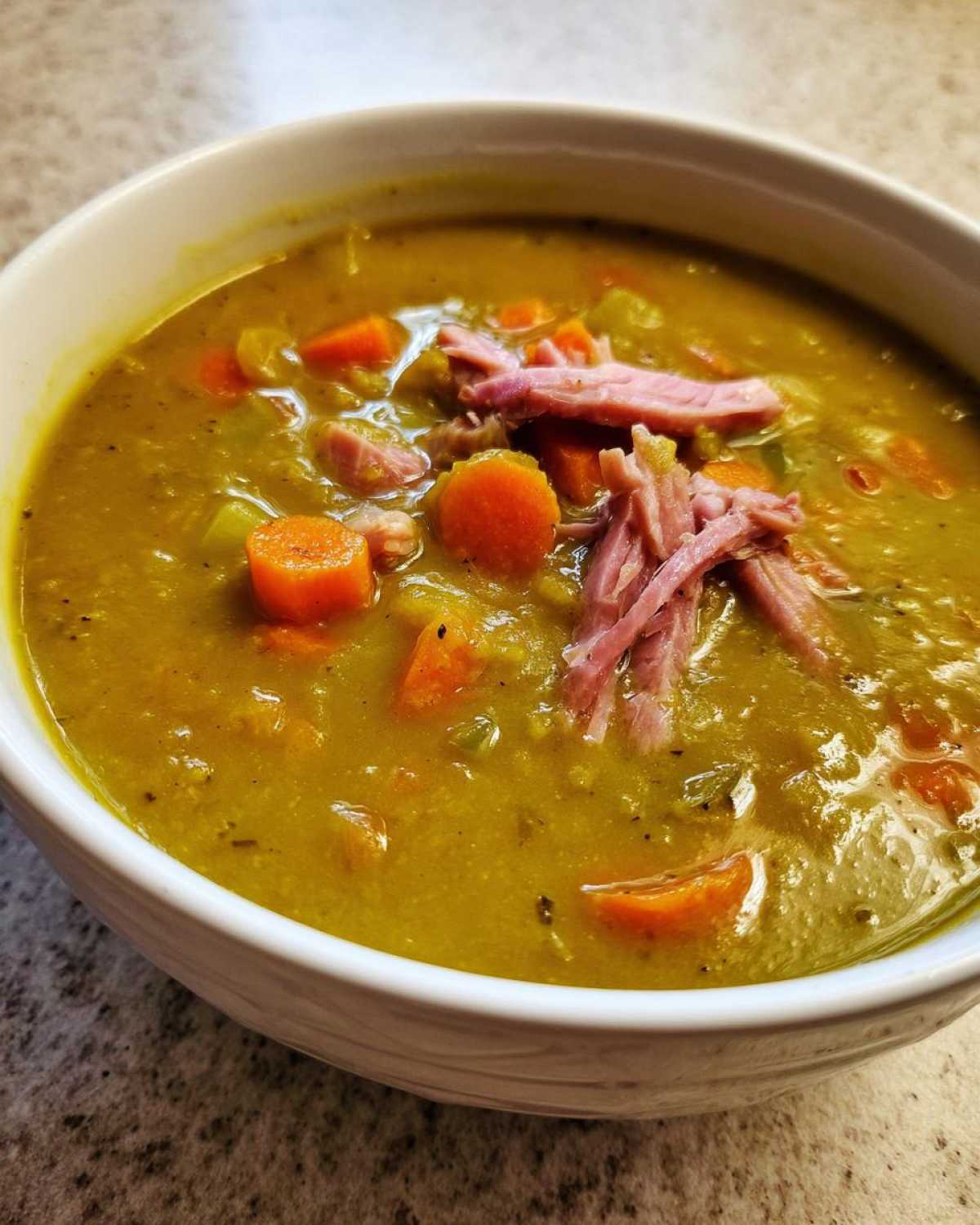 A close-up of a white bowl filled with thick, green pea soup, topped with shredded ham and diced carrots.