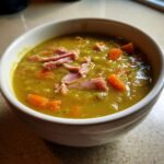 Close-up of a bowl of thick, green pea soup topped with shredded ham pieces and diced carrots.
