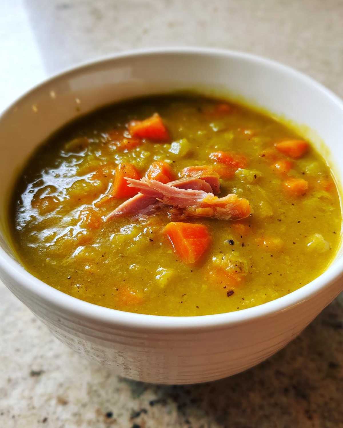 A close-up of a white bowl filled with thick, yellow-green pea soup, topped with shredded ham and diced carrots.