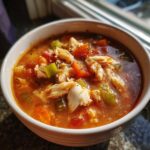 A close-up of a white bowl filled with rich, reddish maryland crab soup, featuring visible chunks of crab meat, carrots, and celery.