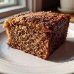 Close-up of a perfectly moist slice of applesauce cake with a dark, sugary crust, sitting on a white plate.