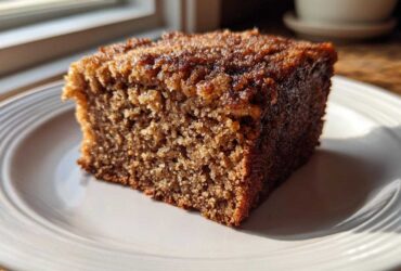 Close-up of a perfectly moist slice of applesauce cake with a dark, sugary crust, sitting on a white plate.