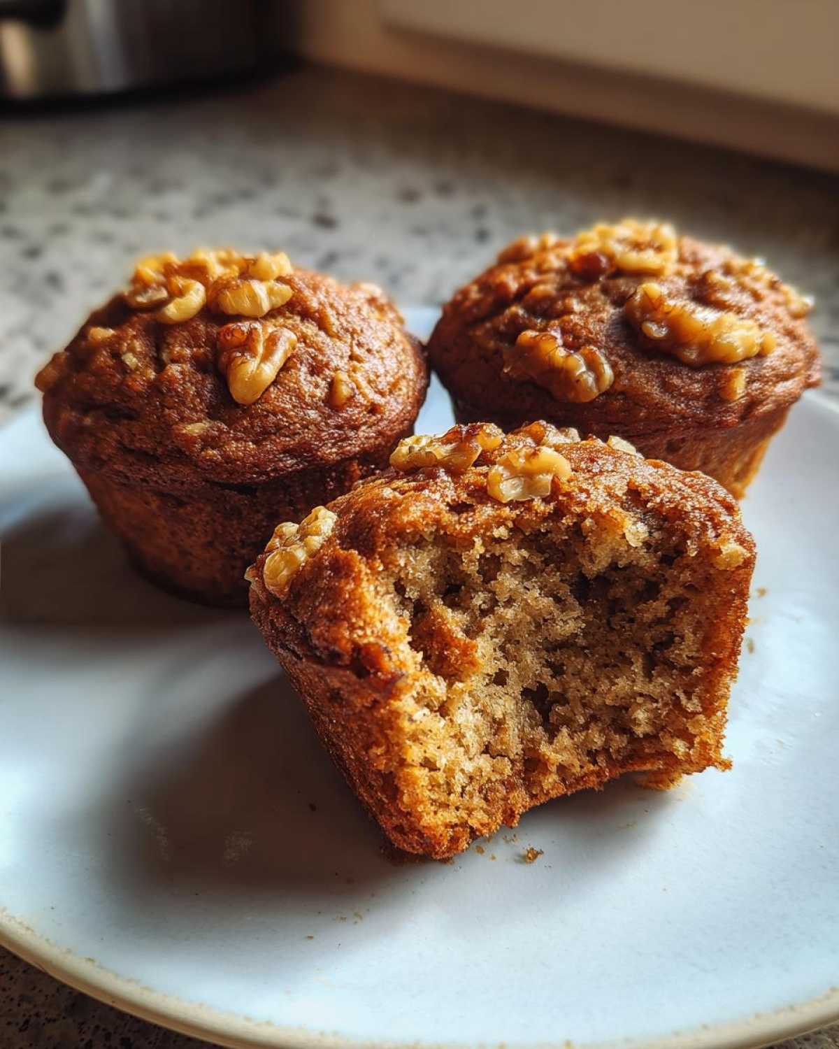 Three moist banana nut muffins on a plate, one bitten to show the texture, topped with walnuts.