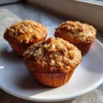 Three golden brown banana nut muffins topped with chopped walnuts resting on a white plate.