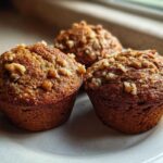 Close-up of three freshly baked, moist banana nut muffins topped with chopped walnuts on a light plate.