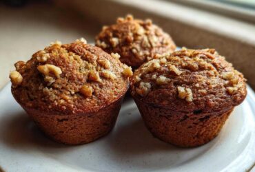Close-up of three freshly baked, moist banana nut muffins topped with chopped walnuts on a light plate.