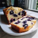 Close-up of a moist blueberry bread loaf with one slice cut and resting next to it on a white plate.