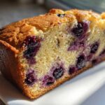 Close-up cross-section of a moist slice of golden blueberry bread packed with dark purple blueberries.