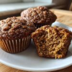 Three freshly baked, dark brown bran muffins on a white plate, one broken open to show the texture.