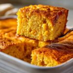 A close-up of a moist, golden slice of cornbread casserole being lifted out of a white baking dish, showing whole corn kernels.