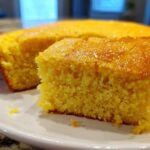 Close-up of a moist, golden slice of sourdough cornbread with a shiny top, resting on a white plate.