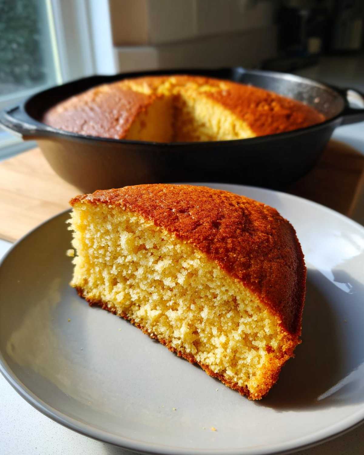 A close-up of a moist slice of golden sourdough cornbread served on a gray plate, with the rest of the cornbread in a cast iron skillet behind it.