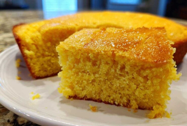 Close-up of a moist, golden slice of sourdough cornbread with a shiny top, resting on a white plate.