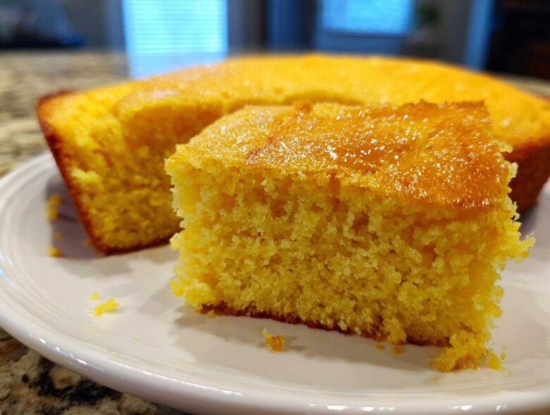 Close-up of a moist, golden slice of sourdough cornbread with a shiny top, resting on a white plate.