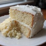 Close-up of a moist slice of vanilla cake recipe layered with white frosting on a white plate.