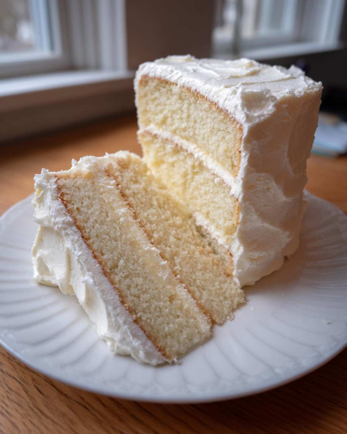 A close-up of two slices of moist vanilla cake recipe layered with thick white buttercream frosting on a white plate.