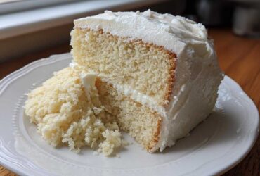 Close-up of a moist slice of vanilla cake recipe layered with white frosting on a white plate.