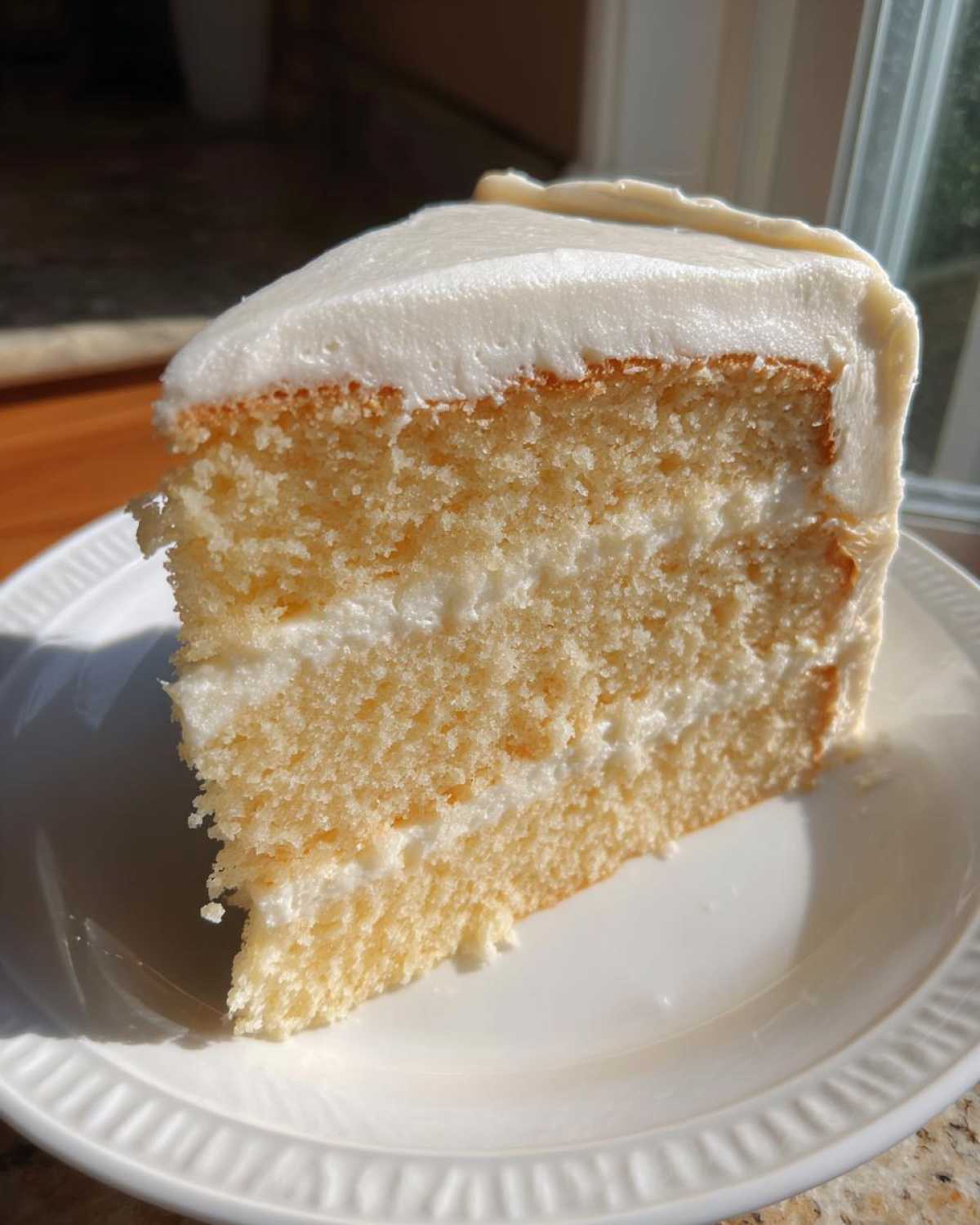Close-up of a moist, two-layer slice of vanilla cake recipe with thick white frosting, sitting on a white plate.
