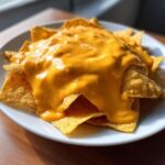 A close-up of tortilla chips completely smothered in thick, bright orange nacho cheese sauce.