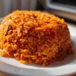 A close-up, featured image of a perfectly molded mound of vibrant orange Nigerian jollof rice on a white plate.