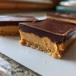 Close-up of two decadent peanut butter bars with a thick chocolate layer on top, showing the crumbly base.