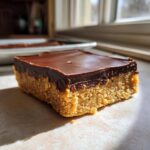 A single, thick square of no bake peanut butter bars featuring a crumbly peanut butter base and a smooth chocolate layer on top.