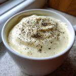 Close-up of creamy Olive Garden Alfredo Sauce in a white bowl, generously topped with cracked black pepper.