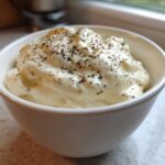 A close-up of thick, creamy Olive Garden Alfredo Sauce topped with freshly cracked black pepper in a small white bowl.