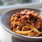 Close-up of fettuccine pasta generously topped with rich, meaty bolognese sauce in a white bowl.