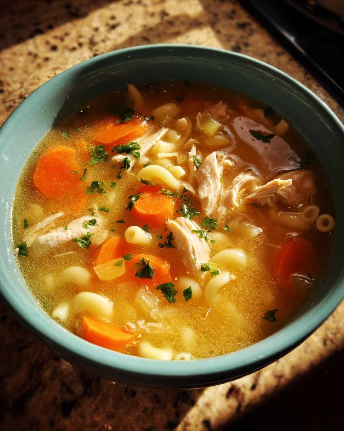 Close-up of a warm bowl of penicillin soup featuring shredded chicken, bright orange carrots, elbow macaroni, and parsley garnish.