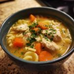 Close-up of a steaming bowl of penicillin soup featuring chunks of chicken, bright orange carrots, and pasta, topped with fresh parsley.