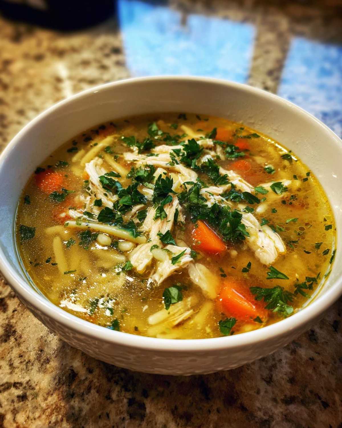 A close-up of a white bowl filled with hot penicillin soup, featuring shredded chicken, carrots, noodles, and fresh parsley.