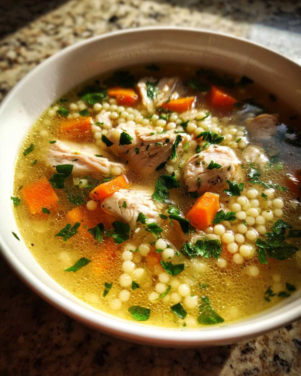 Close-up of a white bowl filled with hot penicillin soup, featuring broth, shredded chicken, small pearl pasta, and diced carrots.