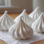 A close-up of several perfectly piped, glossy white meringue cookies resting on crumpled parchment paper.