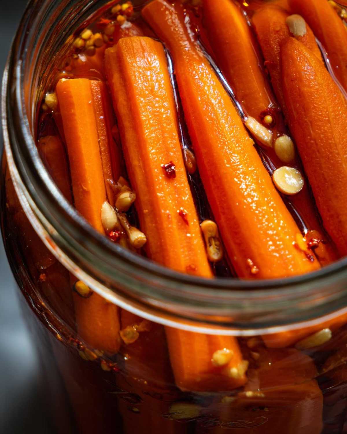 Close-up view of bright orange pickled carrots packed tightly in a glass jar with pickling brine, spices, and seeds.