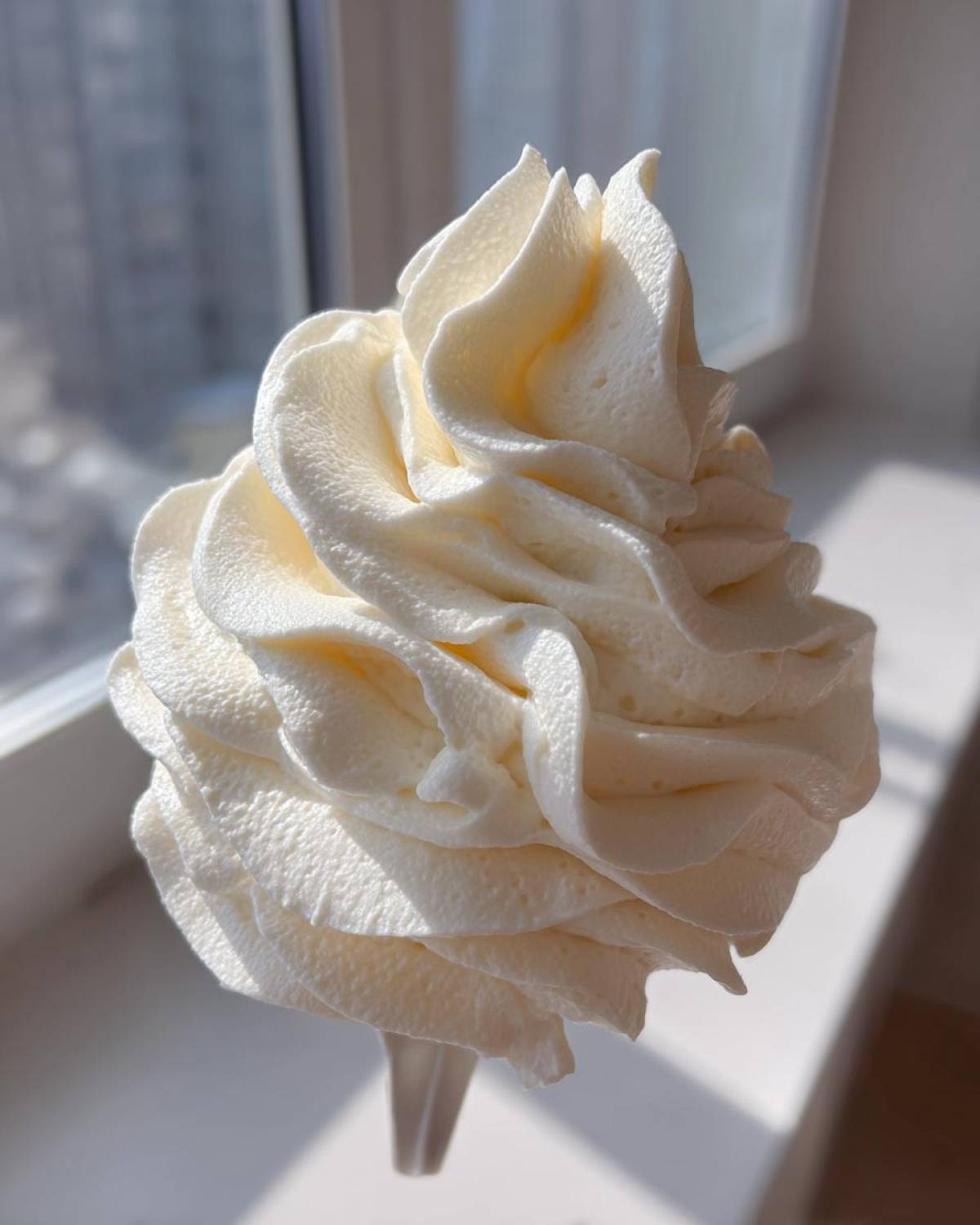 Close-up of a perfectly piped swirl of fluffy, white cream cheese frosting, highlighted by sunlight.