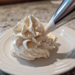 A close-up of a piping bag dispensing fluffy vanilla buttercream frosting onto a white plate.