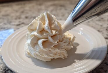 A close-up of a piping bag dispensing fluffy vanilla buttercream frosting onto a white plate.