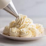 A piping bag with a star tip is used to create swirls of creamy vanilla buttercream frosting on a white plate.