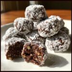 A pile of rich, dark bourbon balls dusted heavily with powdered sugar, one is broken open showing nuts inside.