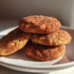 A stack of four soft pumpkin snickerdoodle cookies generously coated in cinnamon sugar, resting on a white plate.