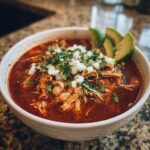 A close-up of a white bowl filled with rich, red chicken pozole, topped with diced onions and cilantro, served with avocado slices.