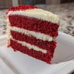 A tall, three-layer slice of vibrant red velvet cake with thick white cream cheese frosting layers.